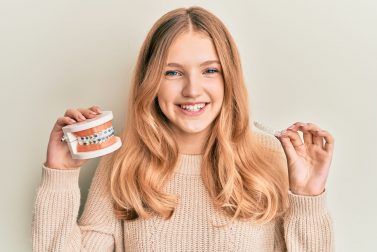 Blonde teenage girl holds up a model mouth with wire braces and clear aligners