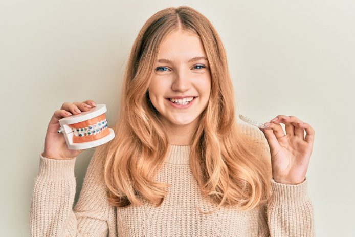 Blonde teenage girl holds up a model mouth with wire braces and clear aligners