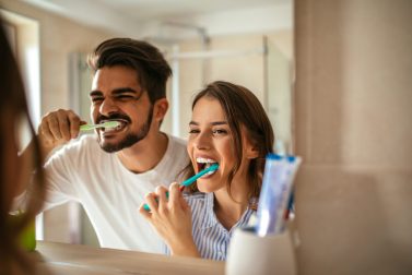 Man and woman smiling and brushing teeth in mirror together